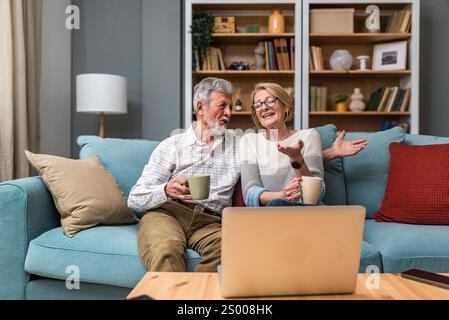 Simple living. Cute senior couple, man and woman, sitting at home on sofa, drinking coffee or tea, watching TV show or movie on laptop computer. Adult Stock Photo
