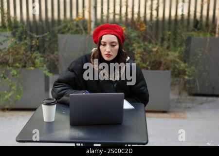 A young woman in a red beret works on her laptop at an outdoor caf, enjoying her coffee Stock Photo