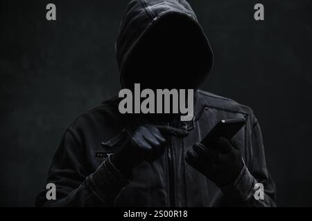 Hooded computer hacker using smartphone, selective focus Stock Photo