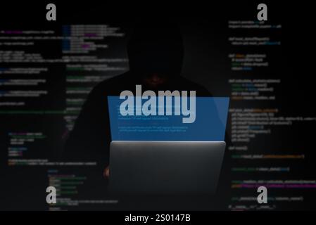Hacker in a dark environment working on a laptop with encrypted code on the screen, symbolizing cybercrime and system breaches. Ideal for cybersecurit Stock Photo