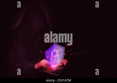 Hooded hacker typing on a laptop with a digital lock shield, symbolizing cyber attacks and data protection IT security. Dark, tech-driven security con Stock Photo