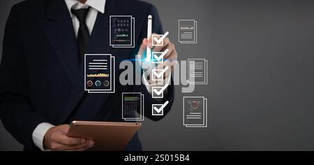 Business professional in a suit uses a digital checklist and tablet to review documents, charts, and data, symbolizing organized project management an Stock Photo