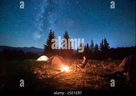 Hiker woman relaxes by campfire, illuminated by its warm glow, with tent nearby under stunning, star-filled sky. Milky Way stretches overhead, adding magical touch to serene mountain setting. Stock Photo