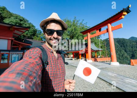 Handsome young tourist enjoying summer holiday in Japan - Traveling life style concept with smiling man taking selfie on city street with japan flag- Stock Photo
