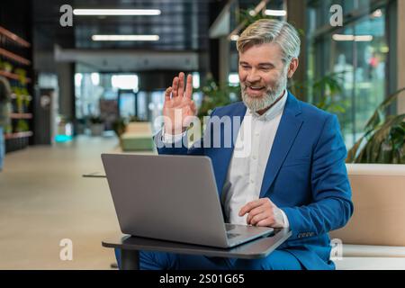 Middle-aged businessman working on laptop computer in modern office lobby talking on online communication video call with employee, boss. Manager man support services looking at webcam of computer. Stock Photo