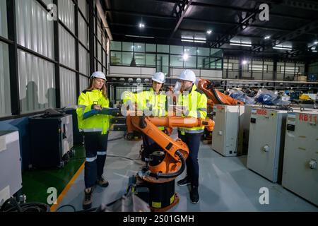 Three engineers in safety gear examine and interact with a robotic arm while discussing its functions inside a well-lit industrial facility. The atmos Stock Photo