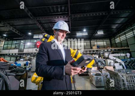 A worker dressed in a suit and hard hat operates a robotic arm in a modern industrial facility. The environment features advanced machinery and a busy Stock Photo