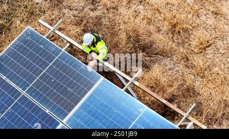 A technician in a safety vest and helmet performs maintenance on solar panels located in an open field, surrounded by dry grass and sunlight. The work Stock Photo