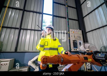 A worker in a yellow safety jacket and hard hat operates a robotic arm in a spacious industrial setting. Sunlight streams through large windows, creat Stock Photo
