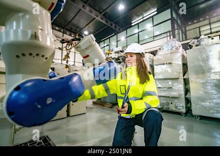 A technician in bright safety clothing reviews a robotic arm in a large warehouse filled with machinery. The setting highlights modern manufacturing t Stock Photo
