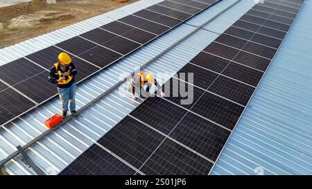 Workers are engaged in the installation of solar panels on a metal rooftop. They wear safety gear and focus on their tasks under clear skies. The sett Stock Photo
