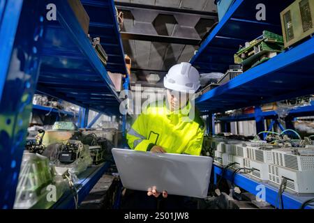 A worker in a reflective yellow jacket and white hard hat focuses on a laptop while surrounded by shelves filled with electronic components. The setti Stock Photo