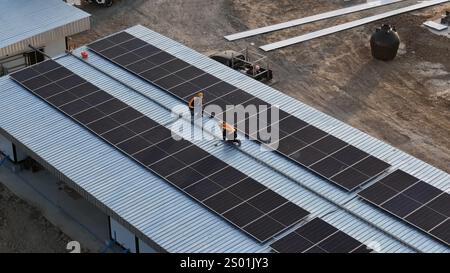 Two workers collaborate to install solar panels on the roof of a commercial building under clear skies. The team's effort contributes to sustainable Stock Photo