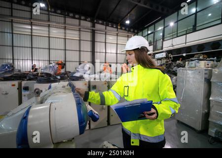 A woman in a bright safety jacket and hard hat examines a robotic arm in an industrial setting. Colleagues work nearby, highlighting a collaborative a Stock Photo