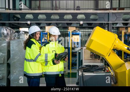 Two workers wearing safety gear are examining robotic equipment in a well-lit industrial facility. They utilize a handheld device to gather data while Stock Photo