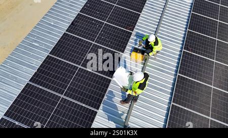 Two technicians in safety gear are working on a rooftop covered with solar panels. One is securing a panel while the other is managing tools and equip Stock Photo