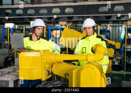 Two female engineers in safety gear inspect robotic arms in a manufacturing facility. One is using a laptop while the other monitors the machinery, fo Stock Photo