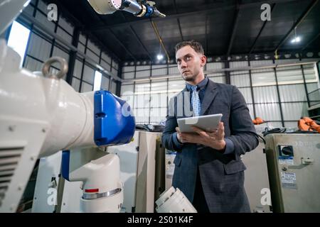 A professional in a suit is observing a robotic arm while holding a tablet. The setting is a large industrial facility with machinery in the backgroun Stock Photo