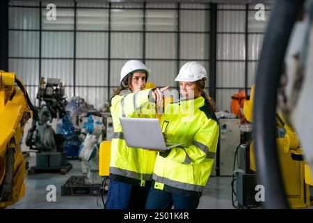 Two engineers in bright safety gear discuss project details while analyzing data on a laptop. The setting is a modern industrial facility with robotic Stock Photo