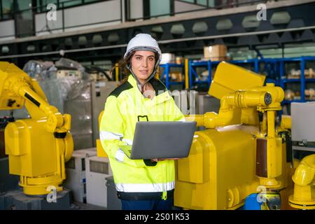 A woman wears a safety helmet and bright jacket while using a laptop in a manufacturing plant. She oversees robotic arms and machinery, ensuring opera Stock Photo