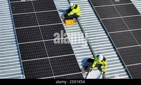 Workers in bright safety vests install solar panels on a metal rooftop under clear skies. Their focus is on aligning and securing the panels for optim Stock Photo