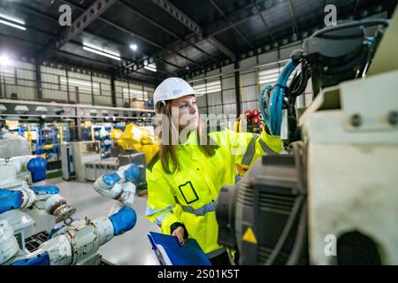 A young female engineer in a safety helmet inspects machinery within a bustling industrial facility. She carefully checks the components, ensuring eve Stock Photo