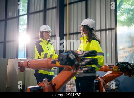 Two engineers in bright safety gear discuss a project while standing next to advanced robotic arms in a manufacturing facility. The atmosphere is focu Stock Photo