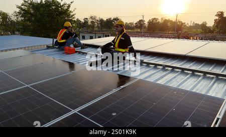 Two workers are engaged in solar panel installation on a rooftop. The sun is setting in the background, illuminating the scene with warm light. Safety Stock Photo