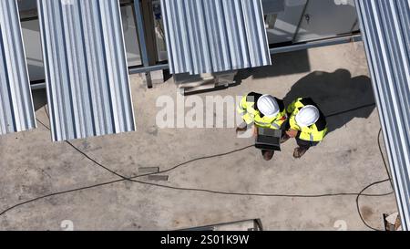 Two construction workers wearing safety helmets and reflective jackets review project plans on a laptop. They are situated on a construction site with Stock Photo