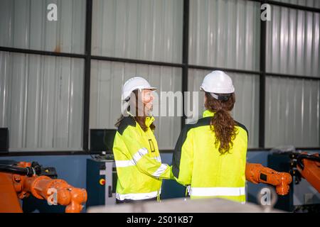 Two engineers wearing safety helmets and reflective jackets discuss their project in an industrial facility. The setting features robotic arms and equ Stock Photo