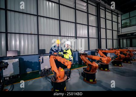 Two engineers discuss robotics systems in a high-tech industrial facility. The workspace is filled with robotic arms, showcasing advanced automation t Stock Photo