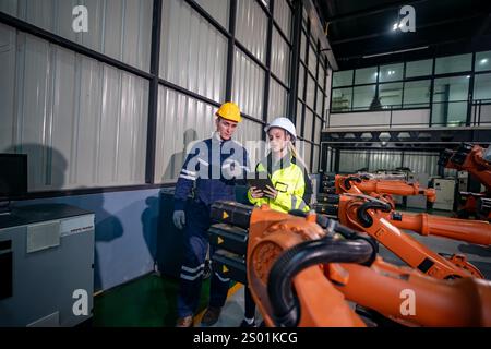 Two engineers collaborate on robotic systems within a well-lit industrial space. They examine data on a tablet while surrounded by advanced machinery Stock Photo