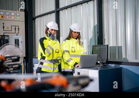 Two engineers wearing safety gear discuss project details in an industrial workspace. One is on the phone while the other operates a laptop at a desk. Stock Photo