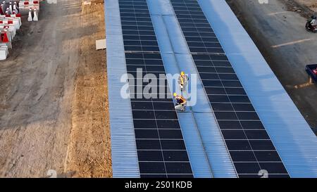 Two workers are installing solar panels on a blue rooftop under clear skies. The construction site is busy with solar panel installation efforts. Stock Photo