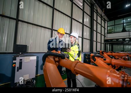 Two engineers discuss robotic systems while examining a tablet in a high-tech factory. They wear safety helmets and work amid advanced machinery, emph Stock Photo