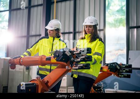 Two individuals in safety gear are focused on controlling a robotic arm in a well-lit industrial space. They are working together, demonstrating teamw Stock Photo