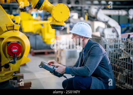 A skilled engineer in a hard hat controls a yellow robotic arm in a busy industrial facility. The expert evaluates machinery and systems while surroun Stock Photo