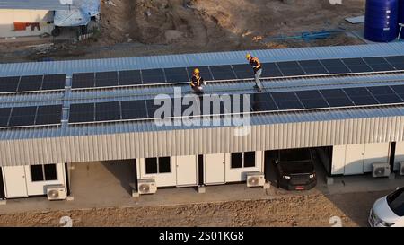 Two workers wearing helmets are seen installing solar panels on the roof of a building under construction in bright daylight. The area shows ongoing w Stock Photo