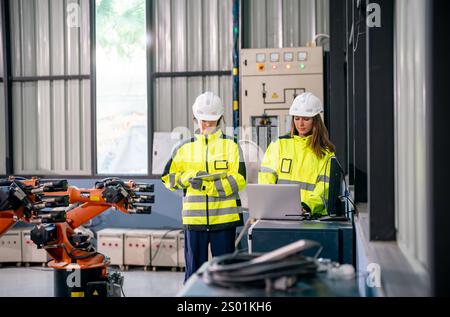 Two workers wearing safety helmets and bright yellow jackets check data on a laptop while overseeing robotic arms in an industrial setting. The enviro Stock Photo