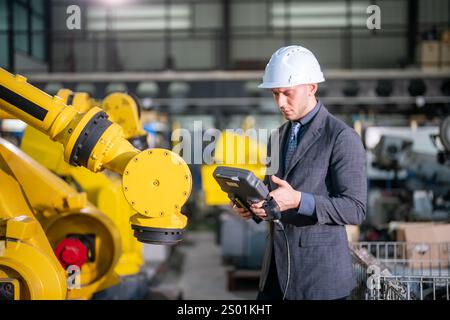 A male engineer in a suit and hard hat uses a handheld device to monitor and control robotic arms in a busy manufacturing plant during daytime operati Stock Photo