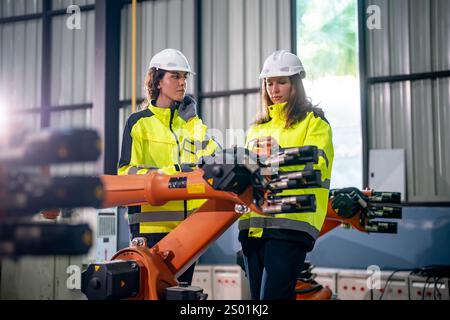 Two female engineers in bright safety gear discuss a robotics project in an industrial setting. They analyze a robotic arm while surrounded by machine Stock Photo