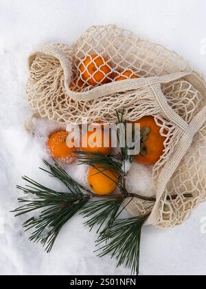 This serene winter photo features bright oranges nestled in a cream-colored net bag placed delicately on a blanket of fresh snow. A green pine branch Stock Photo