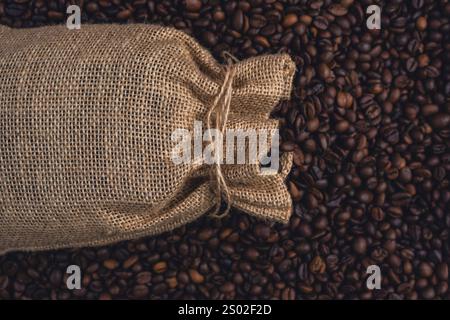 A burlap sack tied with string lying on a bed of roasted coffee beans, representing coffee storage, trade, or rustic aesthetics. Stock Photo