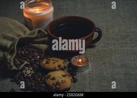A rustic brown ceramic cup of black coffee with chocolate chip cookies, burlap sack spilling coffee beans, and glowing candles on a burlap fabric surf Stock Photo