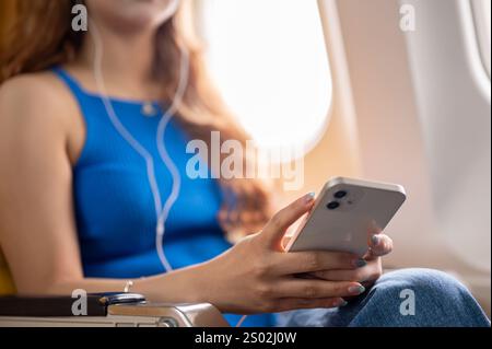 A close-up of a female passenger listening to music through her earphones while using her smartphone during the flight. transport, wireless technology Stock Photo