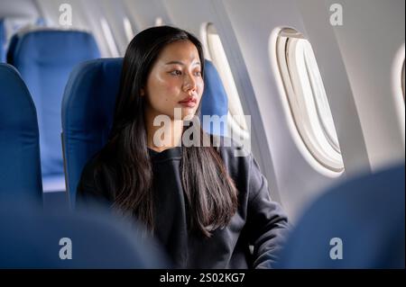 A thoughtful, lonely Asian woman in a cozy black sweater travels alone on holiday, sitting on the flight with a sad expression, gazing out the window. Stock Photo