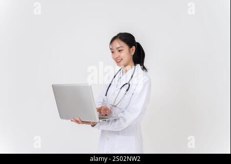 A beautiful, positive Asian female doctor in a uniform stands against a white studio background with her laptop, working on her laptop, typing on the Stock Photo