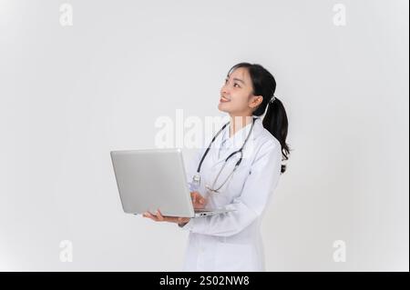 A beautiful Asian female doctor in uniform stands against a white studio background with her laptop, looking upward at an empty space, as she got an i Stock Photo