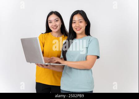 Two happy Asian women with a laptop, standing against a white studio background, smiling at the camera. people and technology concepts Stock Photo
