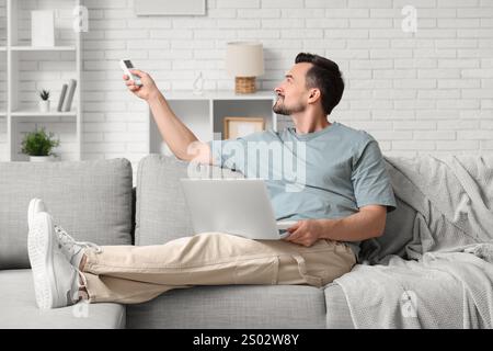 Handsome young man switching on air conditioner while using laptop at home Stock Photo
